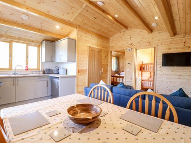 A wooden cabin interior with a kitchen area a dining table with placemats and a wooden bowl blue sofas a TV on the wall and doorways leading to bedrooms at Clover Lodge Theberton near Leiston