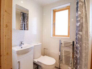 A bathroom with a sink cabinet, toilet, wall mirror, heated towel rail, window, and shower curtain at Clover Lodge in Theberton near Leiston
