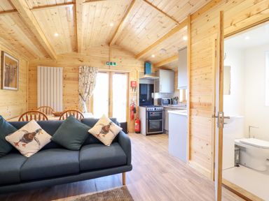 A wooden cabin interior with a blue sofa with duck cushions a dining table a kitchen and a bathroom at Clover Lodge in Theberton near Leiston