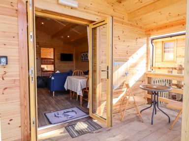 A wooden cabin porch with a small round metal table and two wooden folding chairs at Clover Lodge Theberton near Leiston