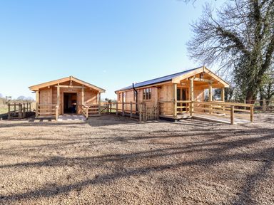 Two wooden cabins with ramps and fences on a gravel ground under clear blue sky at Clover Lodge Theberton near Leiston