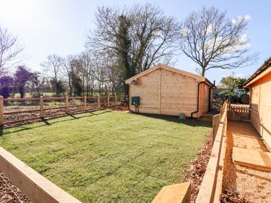 A fenced grass yard with wooden buildings and trees in the background at Clover Lodge in Theberton near Leiston