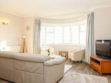 A living room with a beige sofa two white armchairs a wooden TV stand with a television tripod floor lamp wall clock and large windows with curtains at Ocean Watch 5 in Greenhill