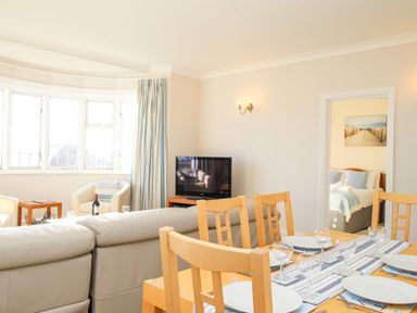 A dining area with a wooden table set with plates and glasses and a living room with a sofa tv and armchairs with a bedroom visible through a doorway at Ocean Watch 5 in Greenhill