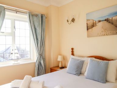 A bedroom with a bed covered in white linens and blue pillows next to a window with curtains and a small wooden nightstand with a lamp and a remote at Ocean Watch 5 in Greenhill