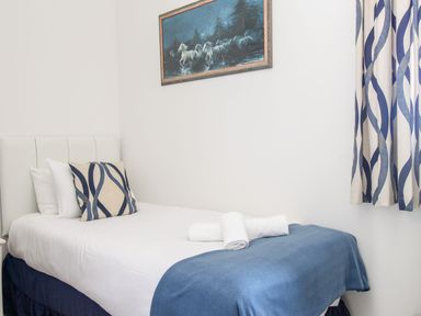 A single bed with white sheets and blue blanket with rolled towels a cushion with blue wavy lines and a painting of horses on the wall in a bedroom at Ocean Watch 5 in Greenhill