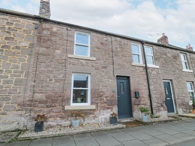 A stone facade house with two windows a dark door and potted plants on a sidewalk at Castle Keep in Norham
