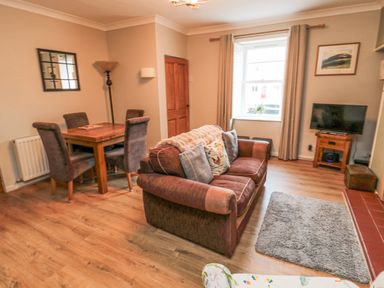 A living room with a brown sofa dining table with four chairs a window with curtains a television on a wooden stand and a floor lamp at Castle Keep in Norham