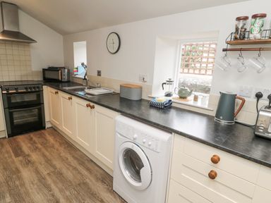 A kitchen with wooden floor a black countertop white cabinets a washing machine a stove a microwave a kettle and cups hanging from a shelf at Castle Keep in Norham