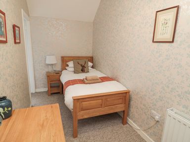 A single bed with folded towels and a cushion with a stag design a bedside table with a lamp framed pictures on the wallpapered walls and a wooden desk at Castle Keep in Norham