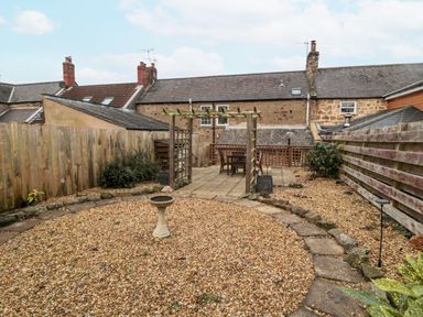 A backyard with a gravel area, stone birdbath, wooden pergola, patio with table and chairs, surrounded by wooden fences at Castle Keep in Norham