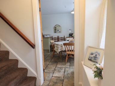 A view through a doorway into a dining room with a table and wooden chairs next to a staircase and a windowsill with a framed picture and flowers at The Old Post House at Low Stott Park near Newby Bridge
