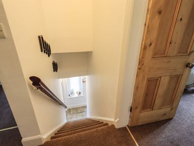 A staircase with brown carpet and wooden handrail leading down past a wooden door to a landing with a window and decorative items at The Old Post House at Low Stott Park near Newby Bridge