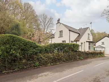 A white house with a stone wall and green hedge beside a road at The Old Post House at Low Stott Park near Newby Bridge
