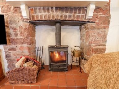 A wood burning stove with fire inside a stone fireplace with a basket of firewood and fireplace tools nearby at Rose Cottage in Holcombe in Teignmouth