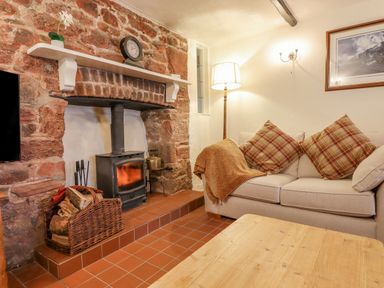 A living room with a wood burning stove in a stone fireplace a basket of firewood a beige sofa with plaid cushions and a blanket a floor lamp and a wooden table at Rose Cottage in Holcombe Teignmouth
