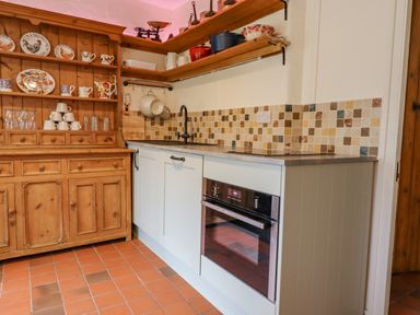 A kitchen with wooden shelves holding plates and mugs and a countertop with a sink and oven at Rose Cottage in Holcombe Teignmouth