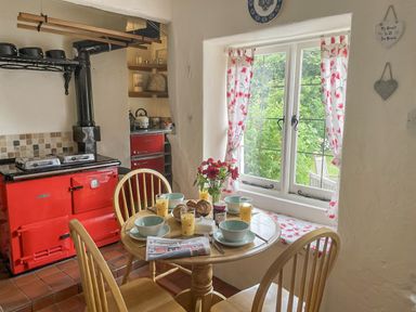 A kitchen with a red stove wooden table and chairs set for breakfast by a window with floral curtains at Rose Cottage in Holcombe Teignmouth