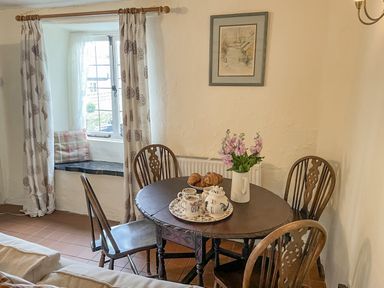 A dining area with a round wooden table with chairs and a window seat with cushions at Rose Cottage in Holcombe Teignmouth