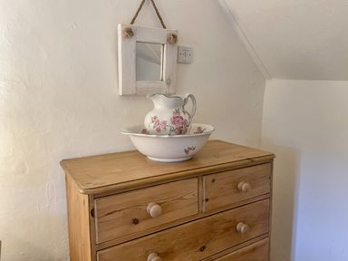 A wooden chest of drawers with a floral pitcher and basin on top and a small square mirror hanging above it at Rose Cottage in Holcombe in Teignmouth