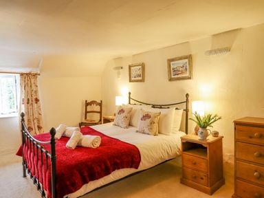 A bedroom with a metal bed frame red blanket wooden furniture and a window with floral curtains at Rose Cottage in Holcombe in Teignmouth