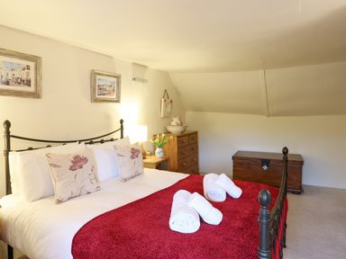 A bedroom with a double bed with red blanket and rolled towels on it next to a wooden chest of drawers and a wooden storage chest at Rose Cottage in Holcombe in Teignmouth
