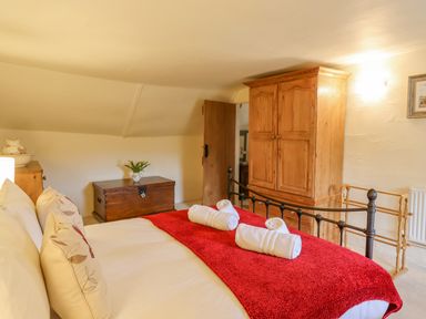 A bedroom with a bed with white sheets and a red throw blanket two rolled towels on the bed a wooden wardrobe a wooden chest with a flower vase and a wooden door at Rose Cottage in Holcombe in Teignmouth