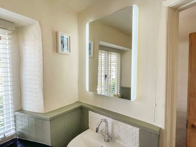 A bathroom sink with a chrome faucet under an illuminated rectangular mirror and a window with blinds at Rose Cottage in Holcombe in Teignmouth