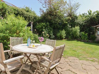 A garden with a round wooden table and four chairs on a stone patio with plants and a small shed at Rose Cottage in Holcombe in Teignmouth