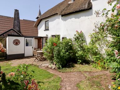 A garden with flowers and a stone patio with wooden chairs and a table outside a thatched roof cottage at Rose Cottage in Holcombe Teignmouth
