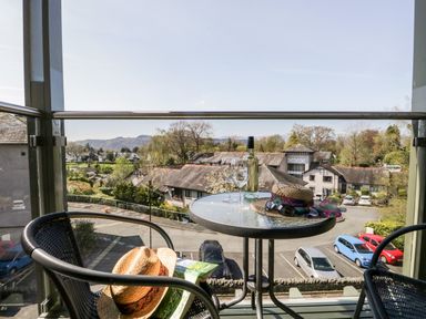 A balcony with a round table holding a bottle of white wine two wine glasses a hat and sunglasses overlooking buildings and parked cars at Claife Heights in Windermere