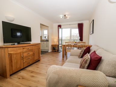 A living room with a beige sofa with pillows a wooden TV stand with a flat screen TV a wooden dining table with chairs and open doors leading to a balcony at Claife Heights in Windermere
