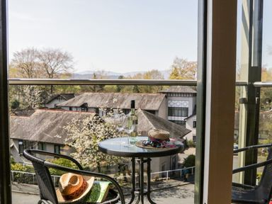 A balcony with a round table holding a bottle, two glasses, a hat, and sunglasses overlooking rooftops and trees at Claife Heights in Windermere