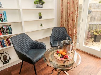Two black chairs near a glass table with champagne bottle in ice bucket and fruit platter in a room with bookshelves and glass door at Calm Skies Scremerston near Berwick-Upon-Tweed