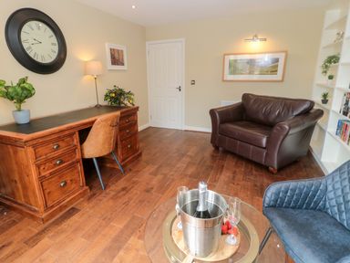 A room with a wooden desk and chair clock on the wall a leather armchair a blue fabric chair a glass table with champagne and strawberries and a white door at Calm Skies Scremerston near Berwick-Upon-Tweed