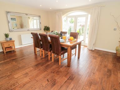 A dining room with a wooden table and six brown chairs next to glass doors with curtains at Calm Skies in Scremerston near Berwick-Upon-Tweed