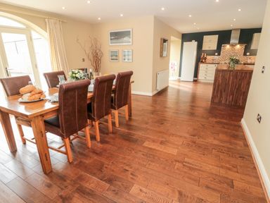 A dining area with a wooden table and six chairs next to a kitchen with wooden cabinets and a white refrigerator at Calm Skies in Scremerston near Berwick-Upon-Tweed