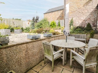 An outdoor patio area with a wooden round table and four chairs surrounded by plants and a stone wall at Calm Skies Scremerston near Berwick-Upon-Tweed