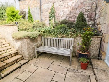 An outdoor patio with stone steps a wooden bench and potted plants against a stone wall at Calm Skies Scremerston near Berwick-Upon-Tweed
