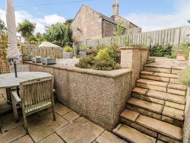 A stone patio with wooden table and chairs next to stone steps leading to a raised garden with planters and bushes at Calm Skies Scremerston near Berwick-Upon-Tweed