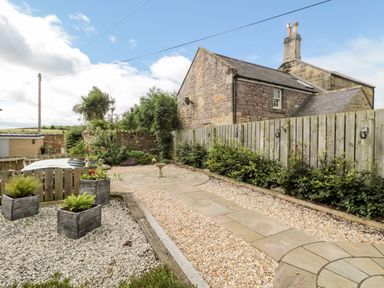 A garden with stone pathways, gravel areas, wooden planters, and a stone house with a chimney at Calm Skies in Scremerston near Berwick-Upon-Tweed