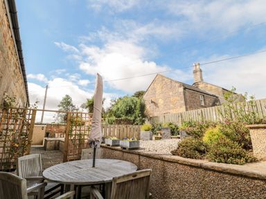 An outdoor patio with a wooden table and chairs a closed umbrella and surrounding plants at Calm Skies Scremerston near Berwick-Upon-Tweed