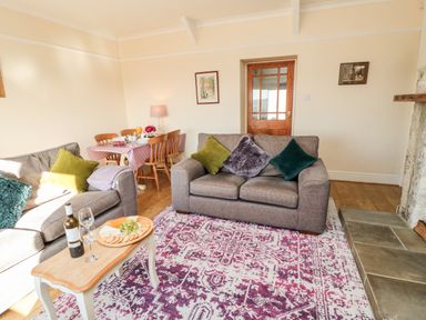 A living room with a patterned rug gray sofas green cushions a wooden coffee table with wine and cheese and a dining table with chairs at Healey Farm Cottage in Rothbury