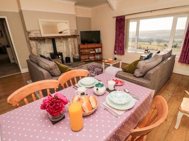 A dining table set with plates cups a teapot and orange juice with a living room with sofas a fireplace and a window with curtains at Healey Farm Cottage in Rothbury