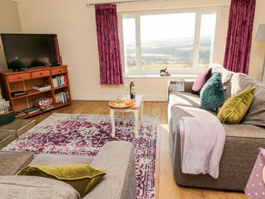 A living room with two sofas a small table with wine and cheese a television on a wooden stand and large windows with purple curtains at Healey Farm Cottage in Rothbury