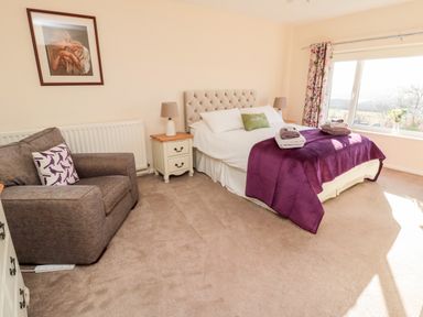 A bedroom with a double bed covered with white bedding and a purple throw blanket beside a window with floral curtains and a gray armchair near a bedside table with a lamp at Healey Farm Cottage in Rothbury
