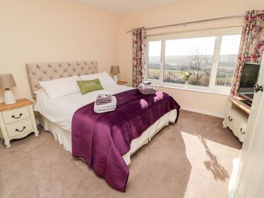 A bedroom with a large bed covered with white and purple bedding two nightstands with lamps a television on a cabinet and a window with floral curtains at Healey Farm Cottage in Rothbury