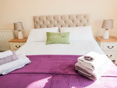 A bedroom with a bed with a green cushion that says Think Happy bee Happy two bedside tables with lamps and folded towels on a purple blanket at Healey Farm Cottage in Rothbury