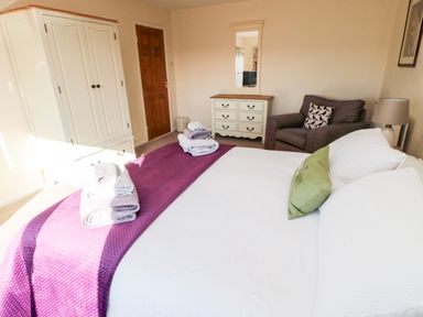 A bedroom with a bed covered in white and purple linens with towels on top a wardrobe a wooden door a dresser with a mirror and an armchair with a pillow at Healey Farm Cottage in Rothbury