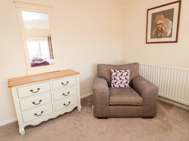 A room with a beige armchair with a pillow beside a white dresser with a mirror and a framed painting on the wall at Healey Farm Cottage in Rothbury
